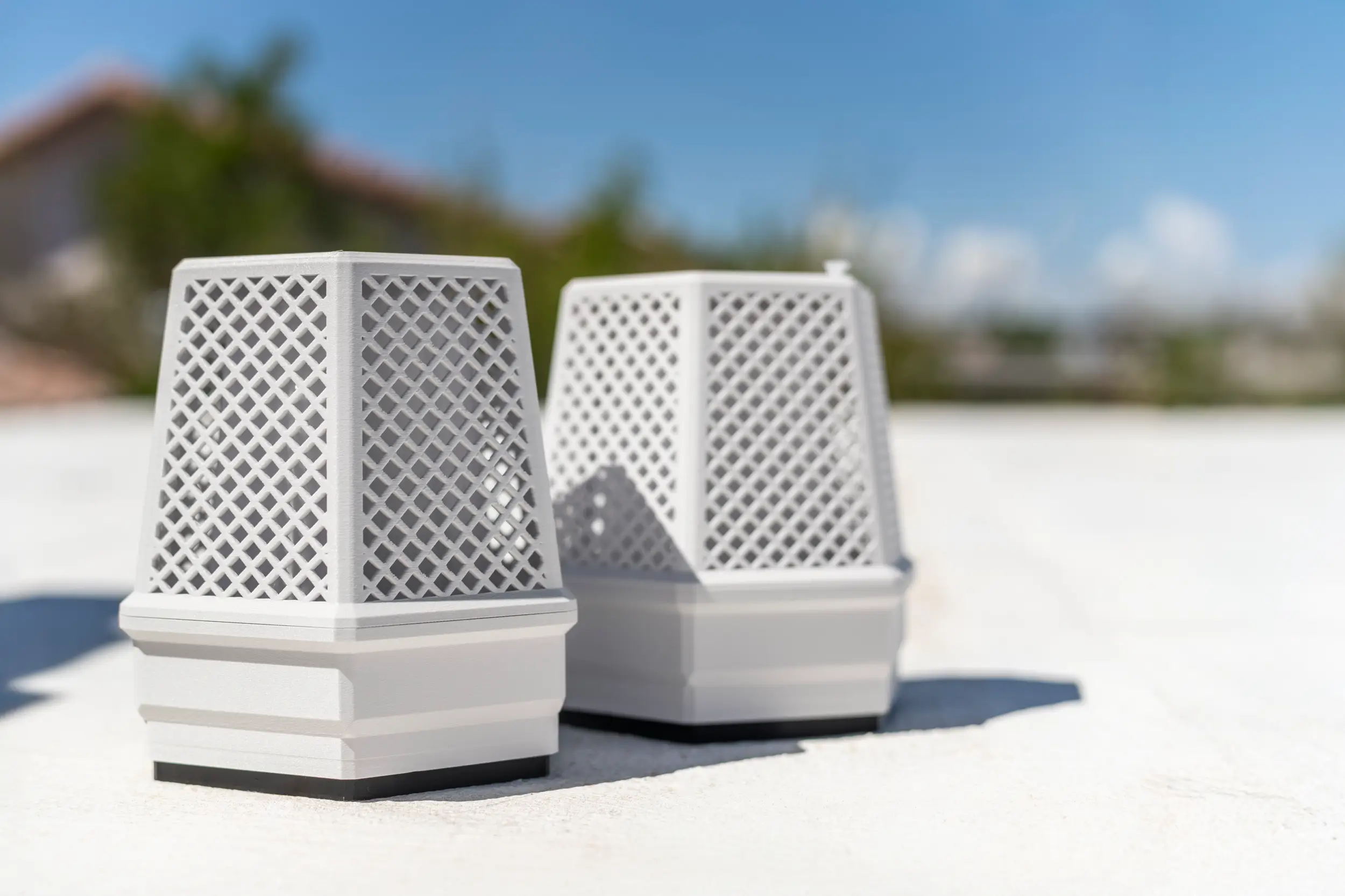 Two white BuzzBox mosquito control units with lattice design, placed outdoors on a bright surface with a blurred house and trees in the background under a blue sky.