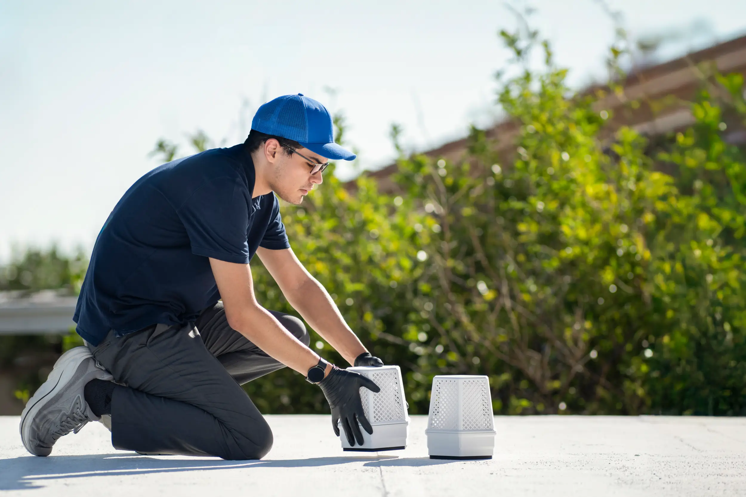 Technician in blue cap and gloves positioning two white BuzzBox devices on a rooftop under bright sunlight.